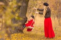 Beautiful mother in a long dress is swinging on a hinged swing little daughter in a red coat in an autumn garden near an old gnarl Royalty Free Stock Photo