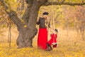 Beautiful mother in a long dress is swinging on a hinged swing little daughter in a red coat in an autumn garden near an old gnarl