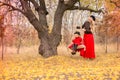 Beautiful mother in a long dress is swinging on a hinged swing little daughter in a red coat in an autumn garden near an old gnarl