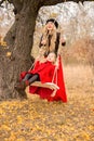 Beautiful mother in a long dress is swinging on a hinged swing little daughter in a red coat in an autumn garden near an old gnarl