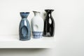 Small decorative porcelain bottles of different colors with drawings and grooves for a better hold placed on a white wooden shelf