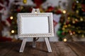 Small easel with framed blank postcard on wooden table with Christmas background.