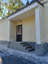 A small entrance with concrete steps leading to a dark wooden door, framed by a simple yellow facade. The scene reflects