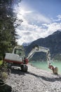 Small excavation digger at gravel shore next to lake, sunny sky