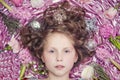 A small fair-haired girl lying on a pink silk fabric with a Christmas garland and Christmas toys around her head