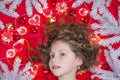 A small fair-haired girl lying on a red floor with a Christmas garland and fir branches around her head