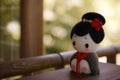 A small figurine of a geisha doll stands against a blurred traditional Japanese background, evoking the elegance and Royalty Free Stock Photo