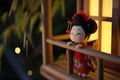 A small figurine of a geisha doll stands against a blurred traditional Japanese background, evoking the elegance and Royalty Free Stock Photo