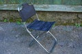 A small folding metal chair with black matter stands on the asphalt against a wall outside