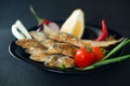 Small fried herrings with vegetables on a black plate on a black background.