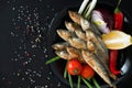 Small fried herrings with vegetables on a black plate on a black background. Fish appetizer.