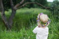 Small girl with blond curly hair wearing colorful wreath made of various wild flowers looking at pine tree. Backview Royalty Free Stock Photo