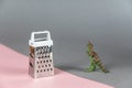 Small grater and dinosaur miniature opposite the gray-pink background. Small green figurine of a predatory dinosaur. Metal grater