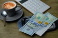 Small gray cup of coffee on a wooden table, metal money tray, american dollars banknotes, Restaurant bill, calculator, tip money Royalty Free Stock Photo