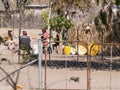 Small group of people sitting around inside fenced compound with their homes and tubs leaning on wall of sticks