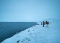 Small group of tourists bundled up on the totally snowy shore of Jokulsarlon lake with icebergs in the background and marveling at