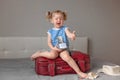 little happy girl sitting burgundy suitcase, holding transparent travel jar full dollars worth money thumbs-up gesture