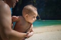 A small infant baby in the arms of dad, very much interested, turned away. Reaches for mom. Toddler on sunny tropical beach