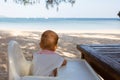 A small infant baby sits in a children chair at a special table and looks at the sandy beach and the sea
