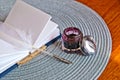 Small journal book and white feather quill lay on a placemat next to an open ink pot