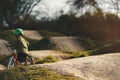 small kid with green helmet rides a balance bike on a dirt jump track in bright sunlight