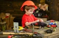 Small kid playing with drill and bolts. Side view boy in orange helmet sitting at table. Little repairman at work