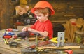 Small kid playing with drill and bolts. Side view boy in orange helmet sitting at table. Little repairman at work