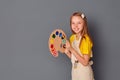 A small laughing girl stands against an empty gray wall with a palette and brushes. She will write a greeting and a drawing. The