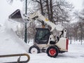 A small loader with a bucket pours snow into a large pile in a city park