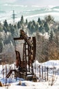 A small metal rusty oil derrick rig on a background of snowy trees and mountains on a sunny day. Vertical orientation.