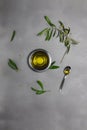 Small metallic bowl and spoon full of olive oil, and cuttings from olive tree with leaves and flowers on grey background