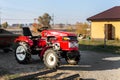 Small mini red modern orange new tractor with trailer standing near hangar building at farm countryside during sunset or