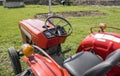 A small mini red tractor stands on a farm yard on green grass and waits for work to begin.