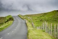 Small narrow asphalt road on a hill between green grass fields. Mountains in low cloud in the background. Connemara, Ireland.