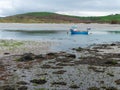 A small old fishing boat is anchored, shallow water at low tide on a cloudy day. Hills under a cloudy sky. Silt