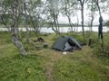 Small open green tent with backpack and hiking gear on grassy shore of Tarra river with birch trees. Swedish Lapland Royalty Free Stock Photo