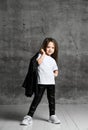 Small positive girl in black and white rock style clothing and white sneakers standing and posing over grey concrete background Royalty Free Stock Photo