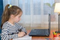 Small preschooler girl in headphones sit at desk study online on laptop, smart little kid wear earphones handwrite in notebook