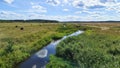 A small river with reflecting clouds flows between an agricultural field and a meadow with cut grass and hay bales. A dirt road ru Royalty Free Stock Photo