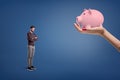 A small sad bearded man stands near a large female hand holding a piggy bank. Royalty Free Stock Photo