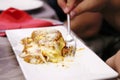 A small serving of lasagne on a square plate, person holds a fork in hand and eats