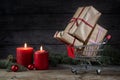 Small shopping cart full with gifts, two red candles, Christmas decoration and fir tree branches on a rustic wooden table, dark Royalty Free Stock Photo