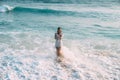 A small slender girl in a beautiful white beach suit is standing in the blue water of the sea, knee-deep. Waves and