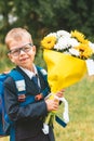 Small smiling boy with flowers ready to go in first class in school. a schoolboy in a school uniform and in a backpack stands on