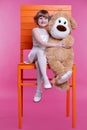 A small smiling girl sits on a huge chair and hugs a large stuffed toy bear Royalty Free Stock Photo