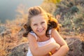 A small smiling laughing girl with curly brown hair dressed white short dress sitting on the stones at the river on Royalty Free Stock Photo