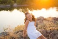 A small smiling girl with curly brown hair dressed white short dress sitting on the stones at the river on sunset time Royalty Free Stock Photo