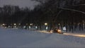 Small snow-removing tractor with headlights turned on cleans the footpath in the city park under the falling snow