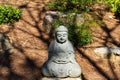 A small stone buddha statue in a Japanese garden surrounded by brown fallen pine needles and lush green trees Royalty Free Stock Photo