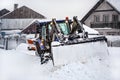 Small tractor with plough removing snow loads during heavy snowstorm calamity, village houses in background Royalty Free Stock Photo